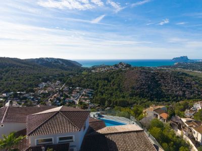 Aerial view showcasing the property's terracotta roofs and pool, nestled amidst green hills, with the Mediterranean Sea and Peñón de Ifach in the distance.