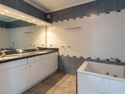 Bathroom featuring a double vanity with dark granite countertop, white cabinets, and an illuminated mirror. Includes a built-in bathtub and grey-white tiled walls.