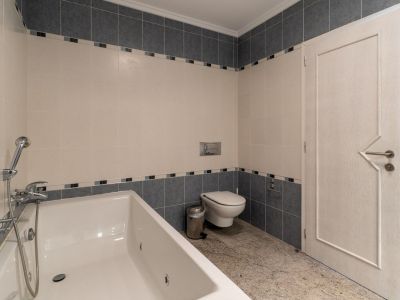 Bathroom featuring a white bathtub with integrated shower, beige and grey tiled walls with a decorative border, and light granite flooring. Includes a wall-mounted toilet and a door with a geometric design.