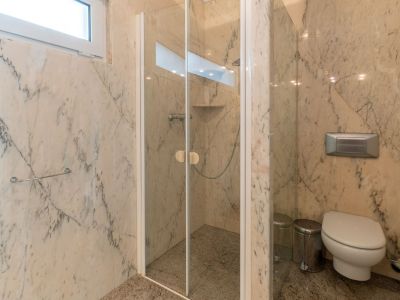 Bathroom featuring veined marble walls and granite flooring. It includes a glass-enclosed shower with an integrated shelf and a wall-mounted toilet, illuminated by a high window.