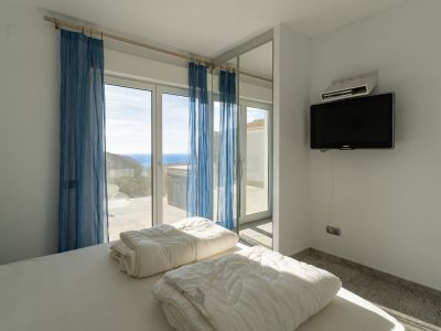 Bedroom featuring a bed with white linen, a wall-mounted TV, and large sliding glass doors with sheer blue curtains, opening to a terrace with panoramic sea views.