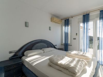 Bedroom with a blue and dark wood double bed, matching bedside tables, and wall lights. Glass doors with sheer blue curtains lead to a terrace, featuring an air conditioning unit.