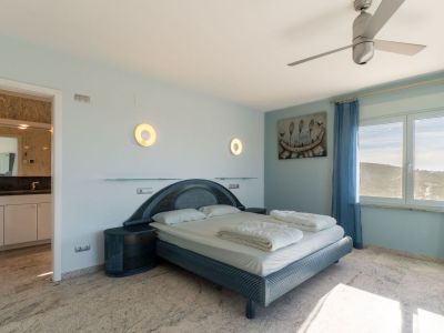 Bedroom with a blue-toned designer bed, matching nightstands, and light marble flooring. The window offers hill views, complemented by blue sheer curtains and an adjacent en-suite bathroom.