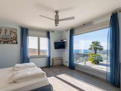 Bright bedroom featuring a large panoramic window with direct sea views and a palm tree. Light granite-effect tiled floor and blue sheer curtains.