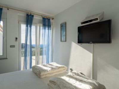Bright bedroom featuring twin beds and direct terrace access. Sea views are visible through sliding glass doors with sheer blue curtains, complemented by a wall-mounted TV.
