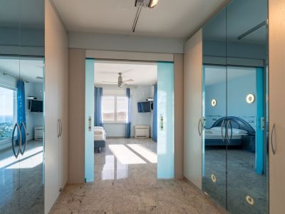 Bright hallway with granite floor and light wood wardrobes. Sliding glass doors frame a bedroom with sea views, while mirrored wardrobe panels reflect another blue-themed bedroom.