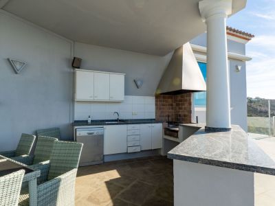 Equipped outdoor kitchen featuring a brick barbecue, granite countertops, sink, and dishwasher, adjacent to a dining table on a covered terrace with hillside views.