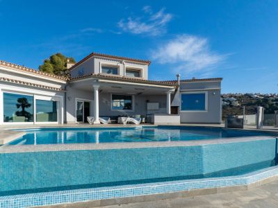 Infinity pool with blue mosaic tiles and covered terrace featuring an outdoor kitchen, adjacent to the villa's facade.