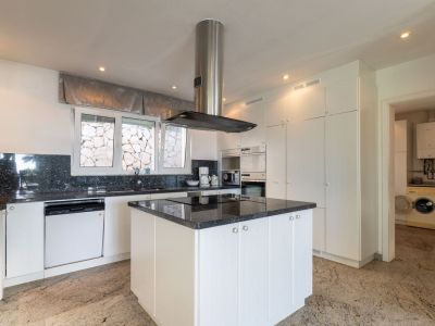 Modern kitchen with white cabinetry, dark granite countertops, and a central island with integrated hob. Integrated appliances, stone wall view from window, and direct access to laundry.