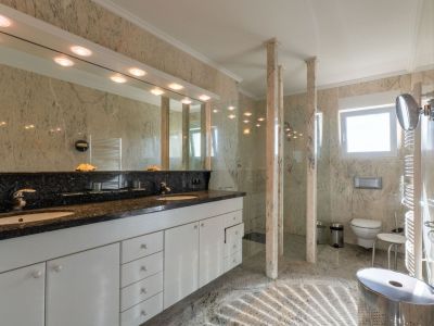Spacious bathroom featuring a double vanity with dark granite countertop and white cabinetry. The walk-in shower with glass partitions and marble pillars complements the light marble walls and floor.