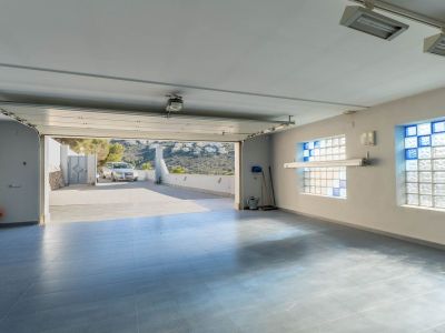 Spacious garage featuring light blue tiled flooring and glass block windows with blue accents, with the main door open to the exterior driveway and distant hills.