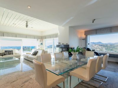 Spacious open-plan living and dining area with polished stone floors. Features a glass dining table, white leather sofas, and a modern fireplace, all bathed in natural light with dual sea and mountain views.