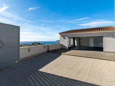 Spacious outdoor area with tiled paving and access to a covered garage. Features panoramic Mediterranean sea and coastal views, framed by a modern grey gate and a low wall with integrated lighting.