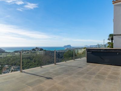 Spacious outdoor terrace with grey tiled flooring and a covered jacuzzi, framed by a glass balustrade showcasing panoramic sea views and the Peñón de Ifach.