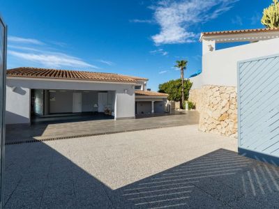 Spacious paved driveway leading to the integrated garage with terracotta tiled roof, framed by a stone wall and a partially open light blue gate under a clear sky.