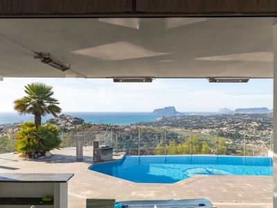 View from inside to the terrace with pool, palm trees, and a vast panorama of the sea, Moraira coastline, and Peñón de Ifach. Ceiling heaters under the overhang.