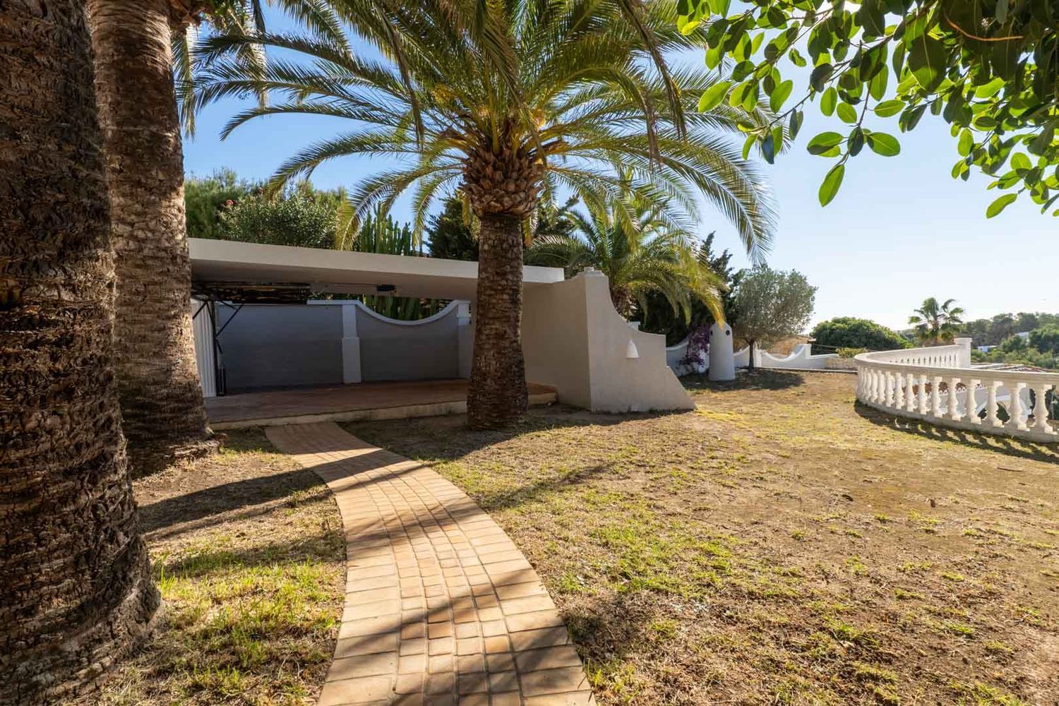 A paved pathway winds through a garden with mature palm trees and green lawn. A covered carport and a white curved balustrade are visible, set against Mediterranean flora under a clear blue sky.