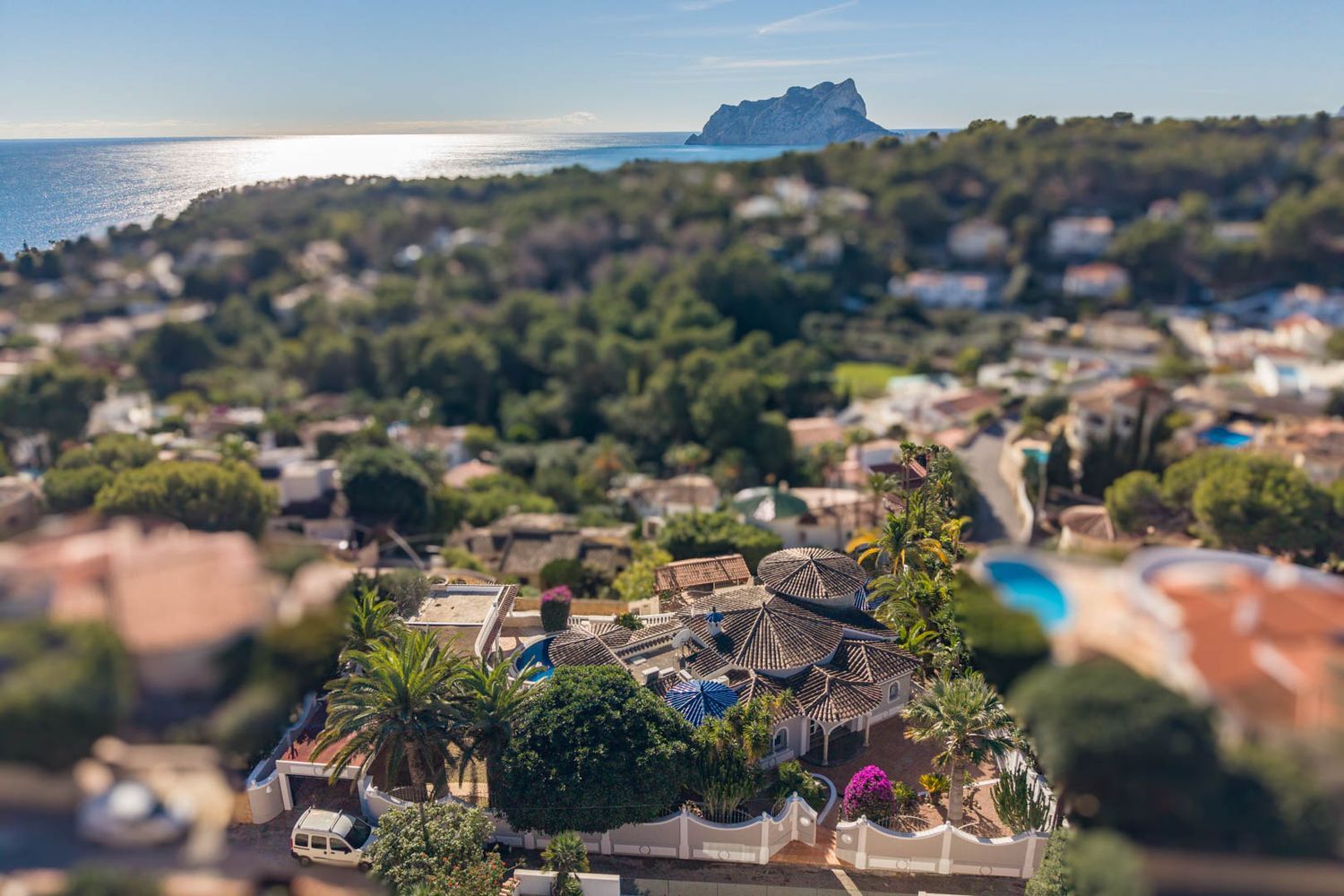 Aerial view showcasing the villa's distinctive tiled roofs amidst lush Mediterranean gardens. The panoramic sea view with the iconic Peñón de Ifach dominates the background.