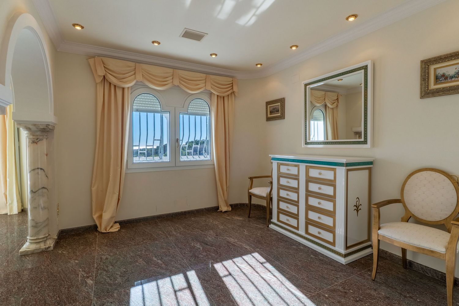 Auxiliary room featuring dark marble flooring, two arched windows with security bars, and beige drapes. A decorative chest of drawers with a mirror and a marble columned archway enhance the space.