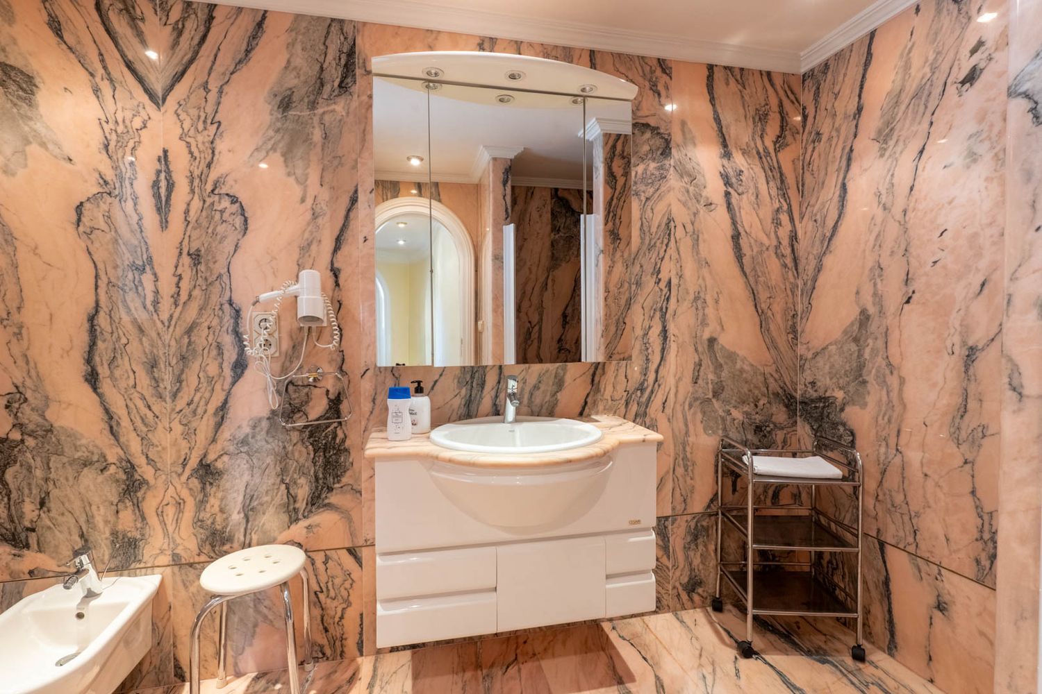 Bathroom clad in striking pink veined marble, featuring a white floating vanity beneath an illuminated mirrored cabinet and a bidet.