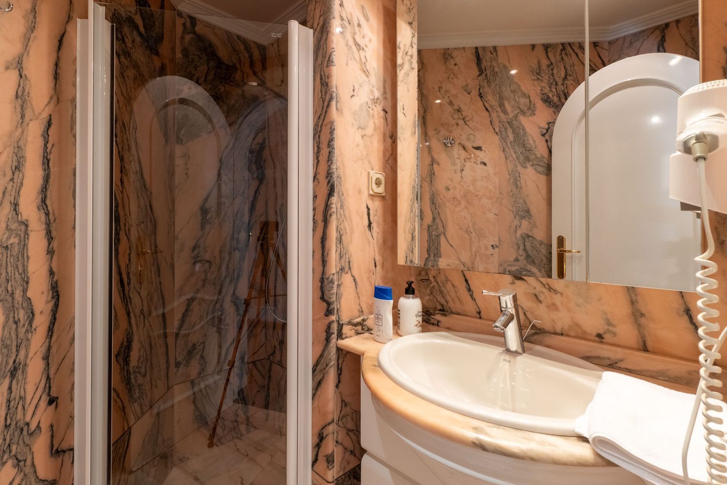 Bathroom featuring striking pink and black veined marble walls, a white ceramic sink, and a glass shower enclosure with a chrome faucet.