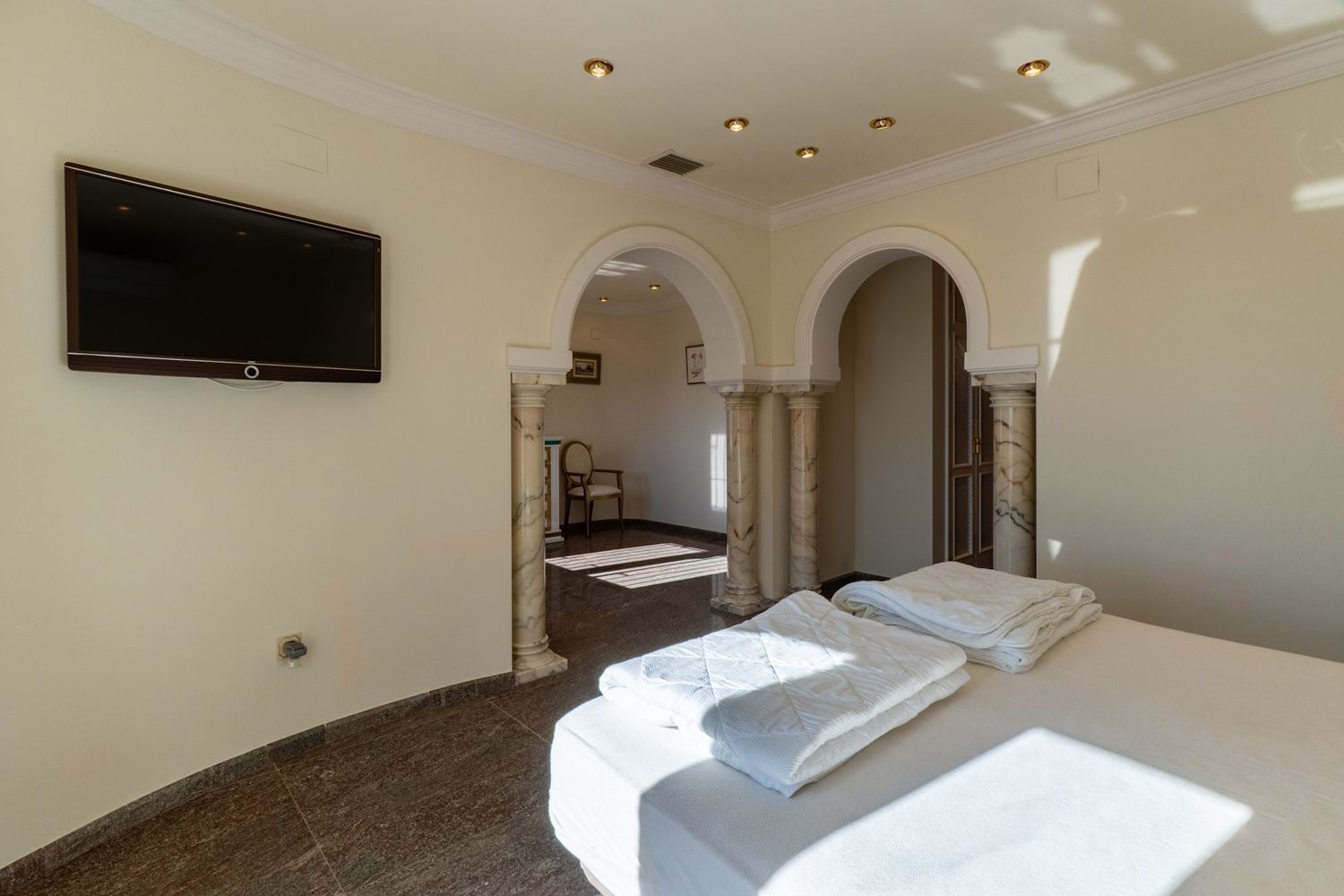 Bedroom featuring a bed, dark marble flooring, and a wall-mounted TV. Two arched passages with decorative marble columns open to an adjacent room, illuminated by recessed lights and natural sunlight.