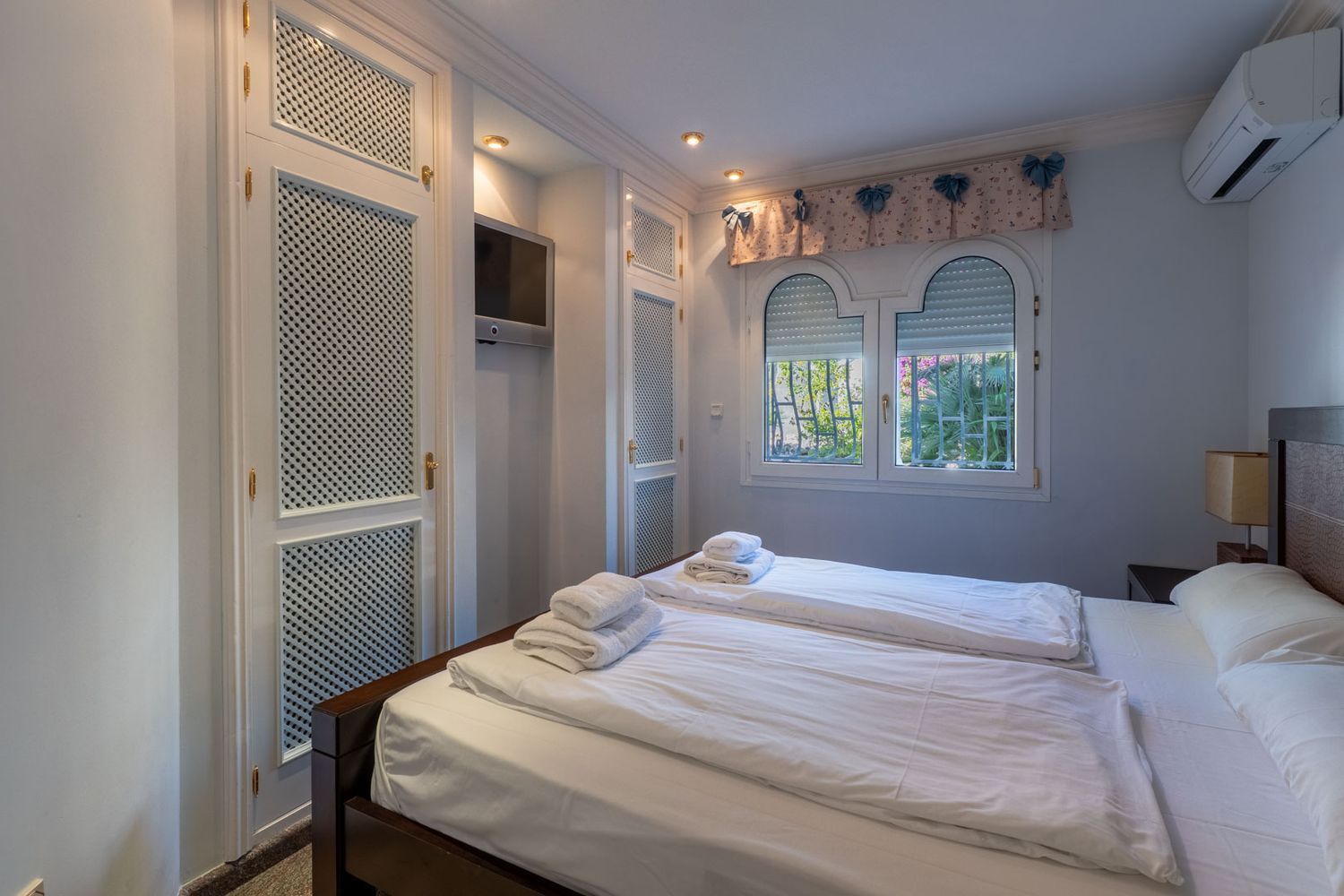 Bedroom featuring white louvered built-in wardrobes, a wall-mounted TV, and arched windows with garden views.