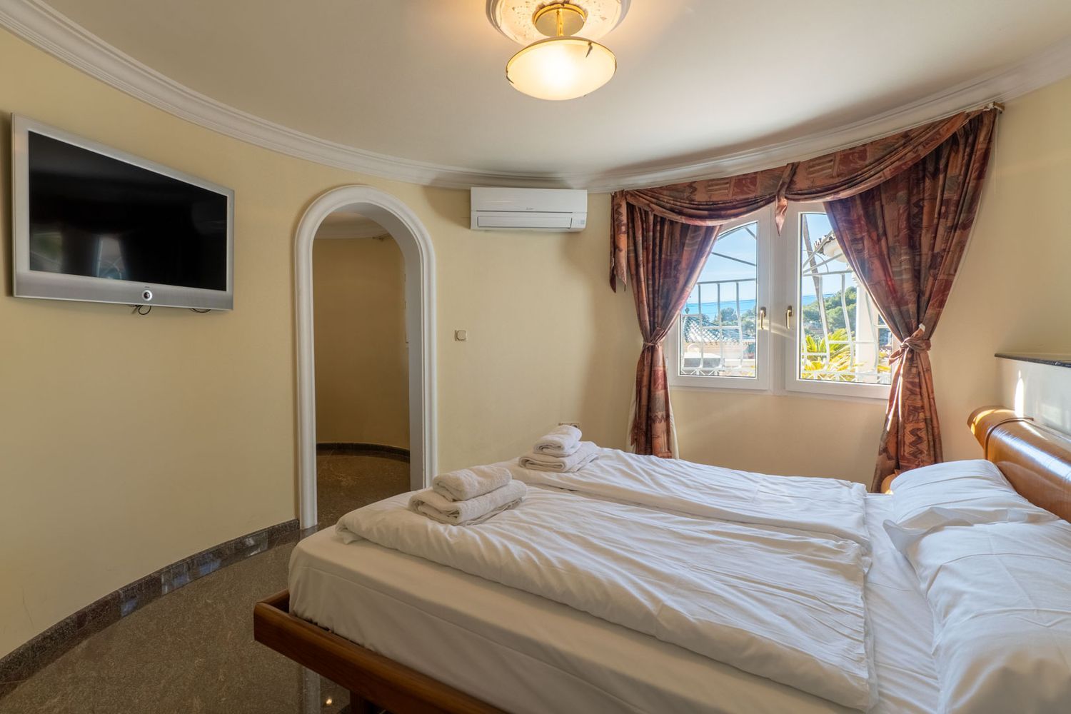Bright bedroom featuring twin beds, a wall-mounted TV, and partial sea views through windows with patterned curtains. Includes air conditioning and an arched doorway.
