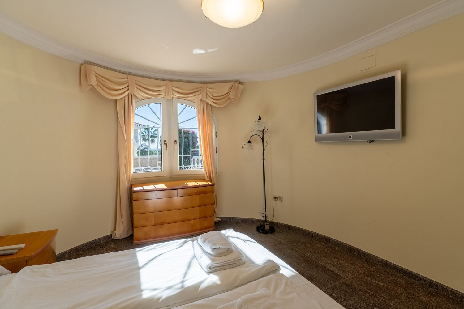 Bright bedroom with a bed, light wood chest of drawers under a curved window, and wall-mounted TV. Dark tiled floor and light walls are bathed in natural light, with a glimpse of outdoor greenery.