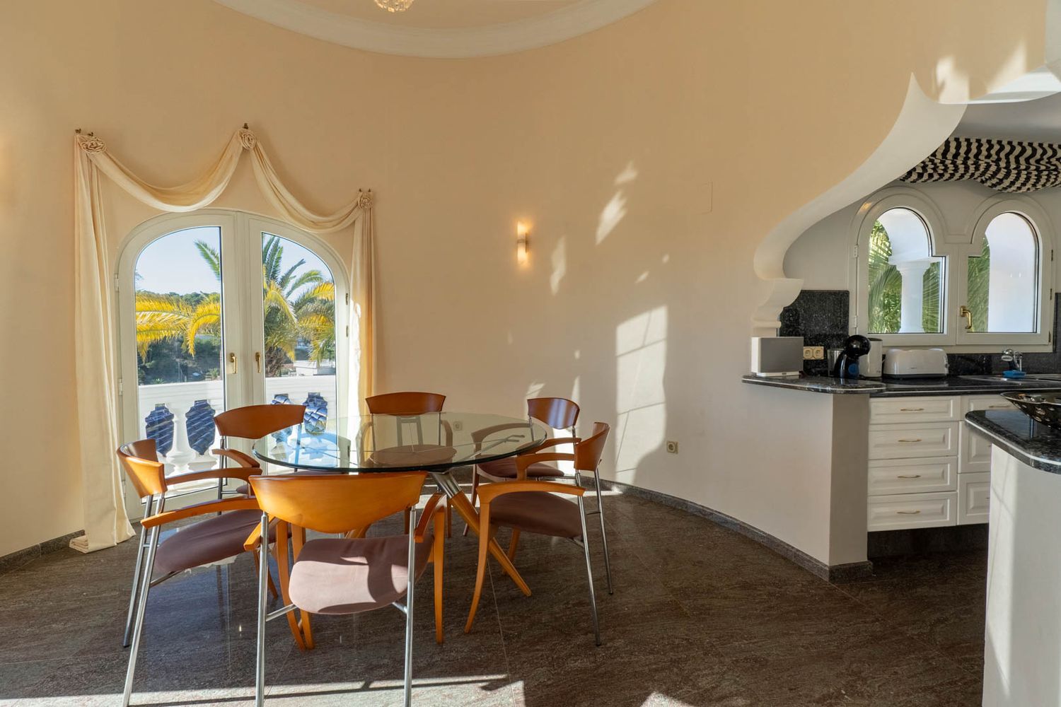 Bright dining area featuring a glass table and wooden chairs, connected to a kitchen bar with arched windows, and offering views of palm trees through French doors.