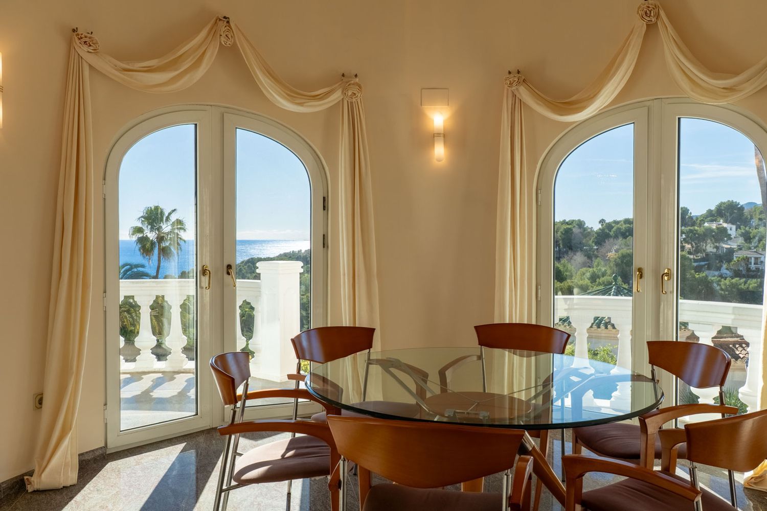 Bright dining area featuring a glass table and wooden chairs, framed by arched doors opening to panoramic Mediterranean sea and coastal views.