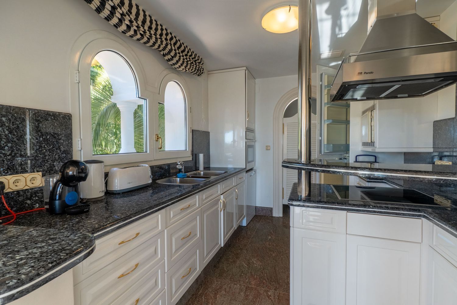 Bright kitchen featuring dark granite countertops, white cabinetry, and arched windows overlooking lush greenery. Equipped with a breakfast bar and modern appliances.