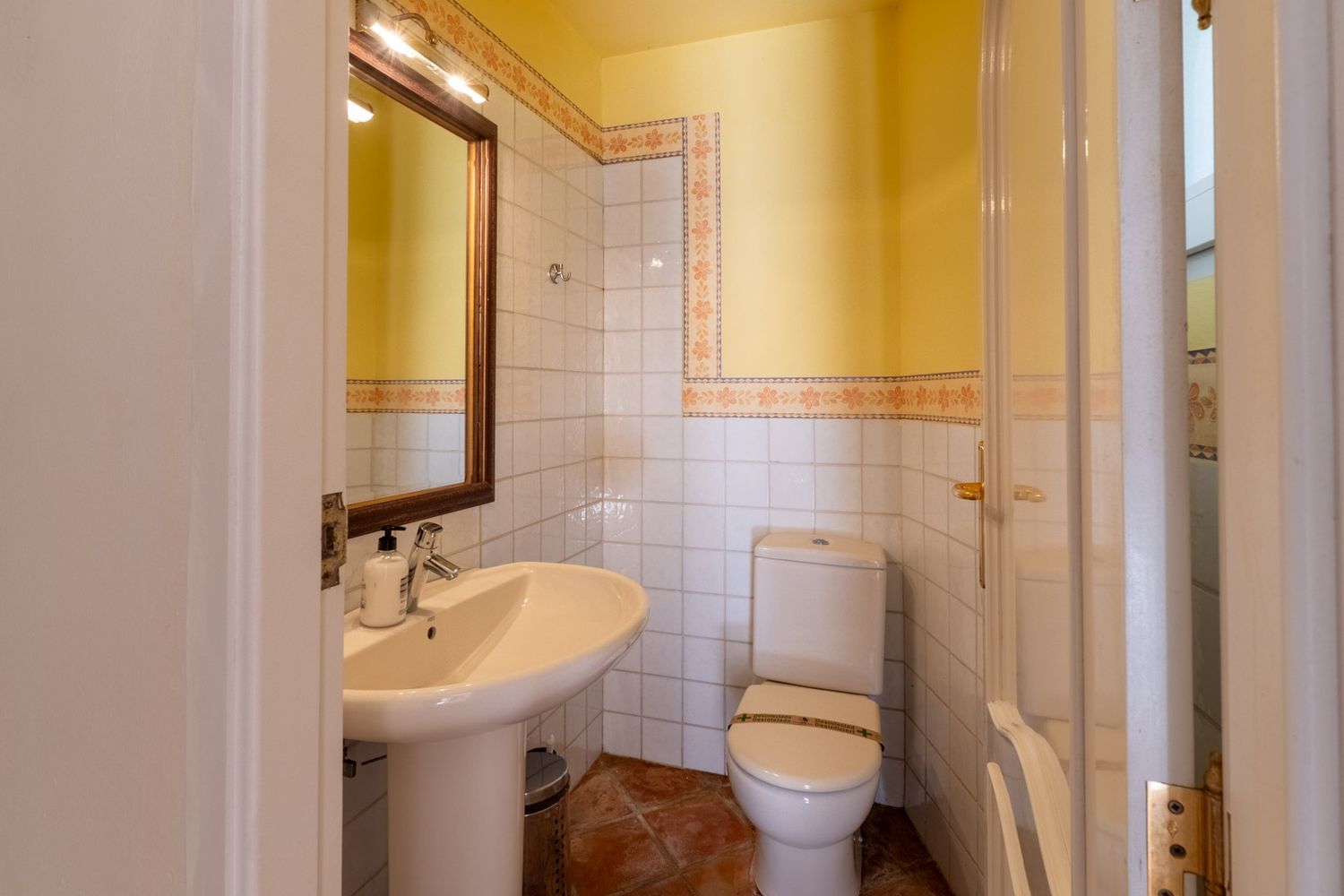 Compact bathroom featuring a pedestal sink, a wooden-framed mirror with overhead lighting, and white tiled walls with decorative floral borders. It includes a toilet and terracotta floor tiles.