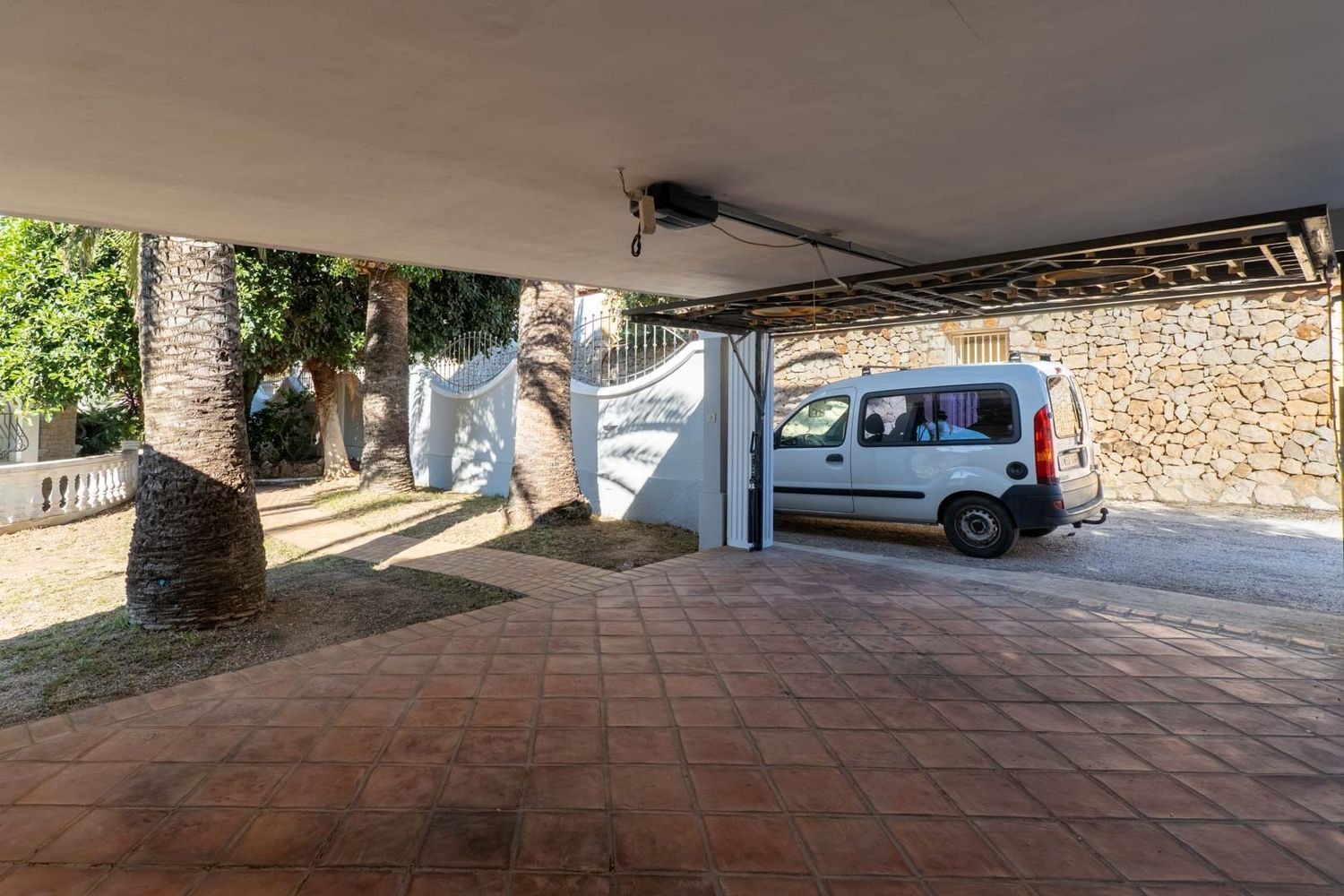 Covered parking area with terracotta tiled flooring, bordered by mature palm trees and a white van parked. A stone wall is visible in the background.