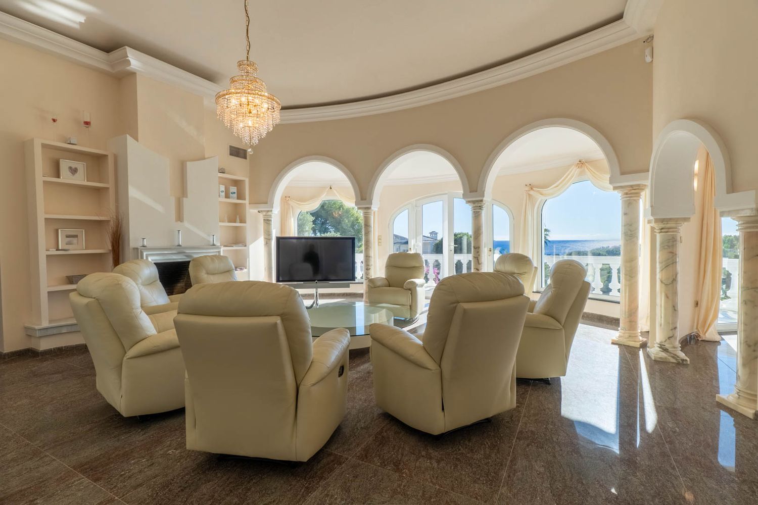 Curved living room featuring recliners, polished marble flooring, and sea views through arched windows. A central crystal chandelier and classic columns enhance the elegant space.