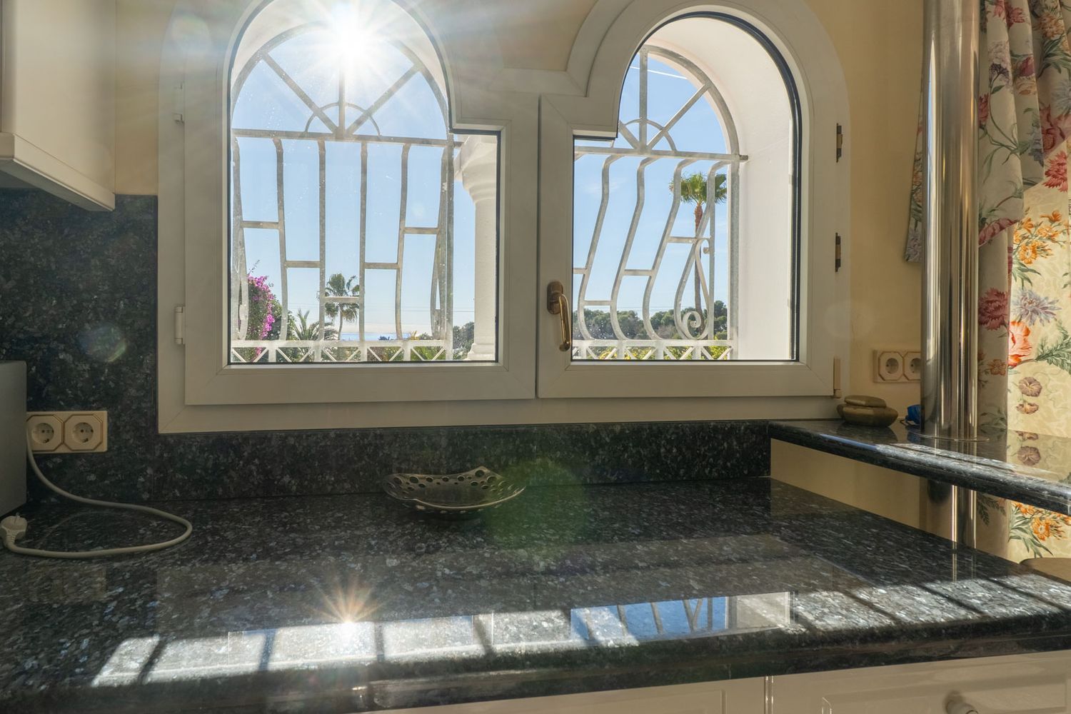 Dark granite countertop beneath two arched windows with decorative grilles, revealing partial sea and palm tree views. Direct sunlight illuminates the surface.