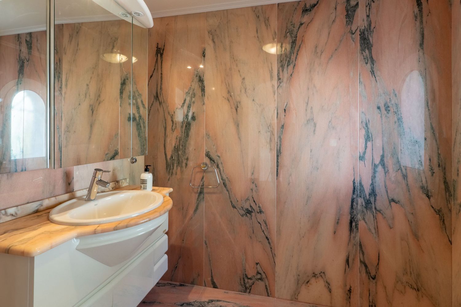 Elegant bathroom featuring walls and countertop clad in distinctive pink marble with dark veining. A white vanity with an oval sink and mirrored cabinet provides functionality.