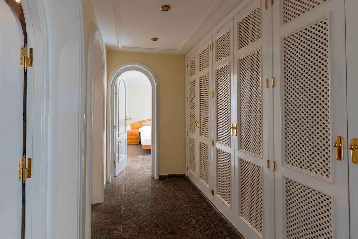 Elegant hallway featuring extensive white louvered built-in wardrobes with brass handles, dark tiled flooring, and arched doorways leading to bright rooms, including a bedroom.
