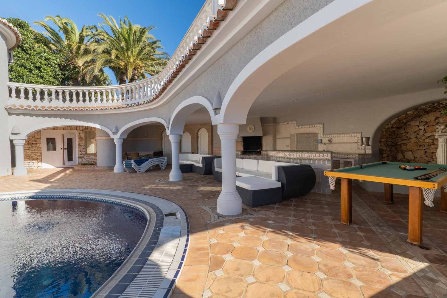 Expansive covered terrace featuring a curved pool, outdoor kitchen with barbecue, lounge seating, and a billiard table on terracotta flooring.