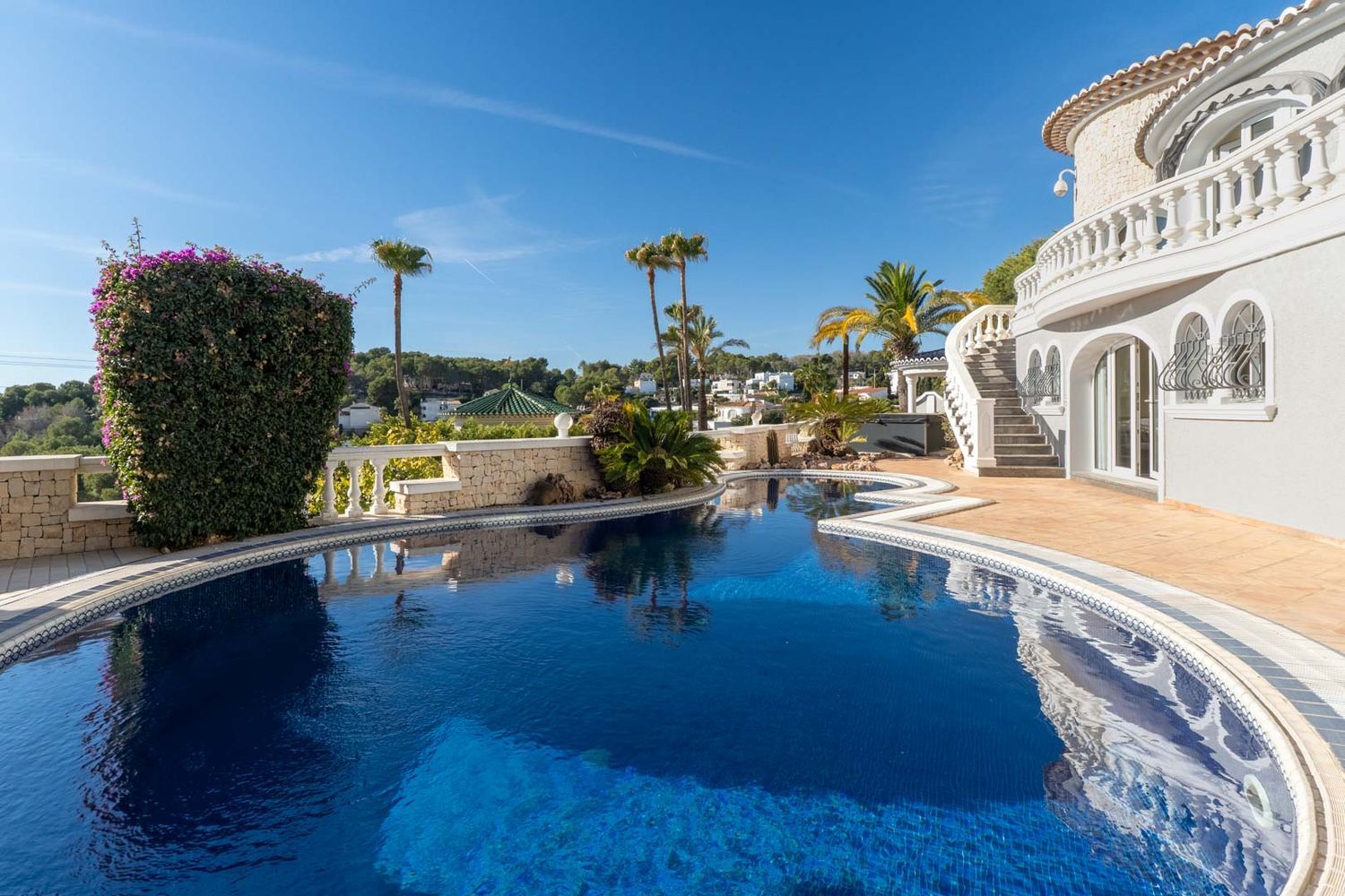 Expansive private pool with blue mosaic tiles and an irregular shape, surrounded by light paving and Mediterranean flora. A grand curved staircase with white balustrades leads to the villa.