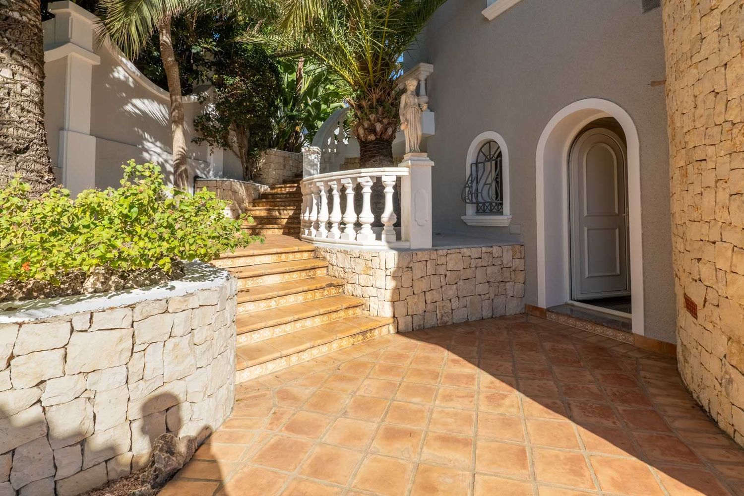 Exterior entrance featuring terracotta tiled flooring and ascending steps. A classical white balustrade and natural stone walls frame the grey facade with an arched doorway.