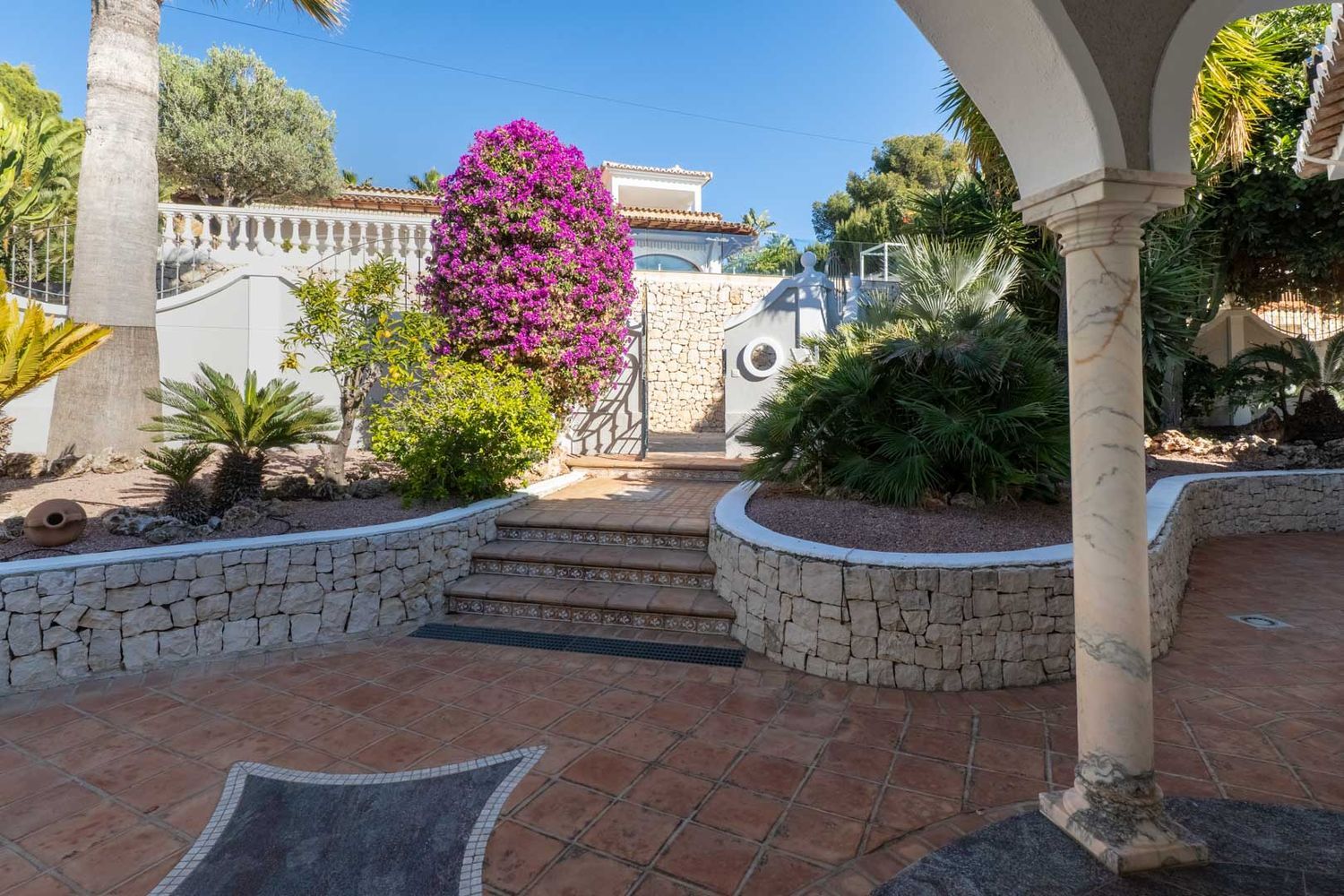 Exterior patio featuring terracotta tiles with a dark mosaic inlay, leading to decorative steps, a stone wall, and a gate adorned with vibrant purple bougainvillea. A marble column frames the view.