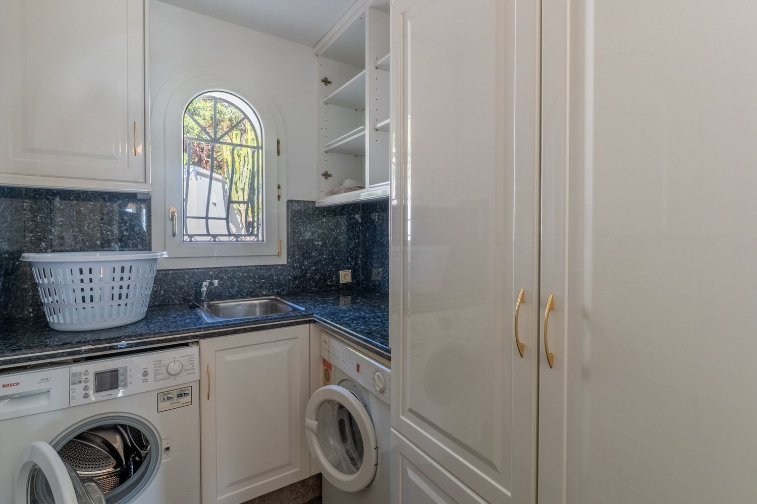 Functional utility room featuring dark granite countertops, a stainless steel sink, and washing machines. An arched window with a grate provides natural light, complemented by white cabinetry.