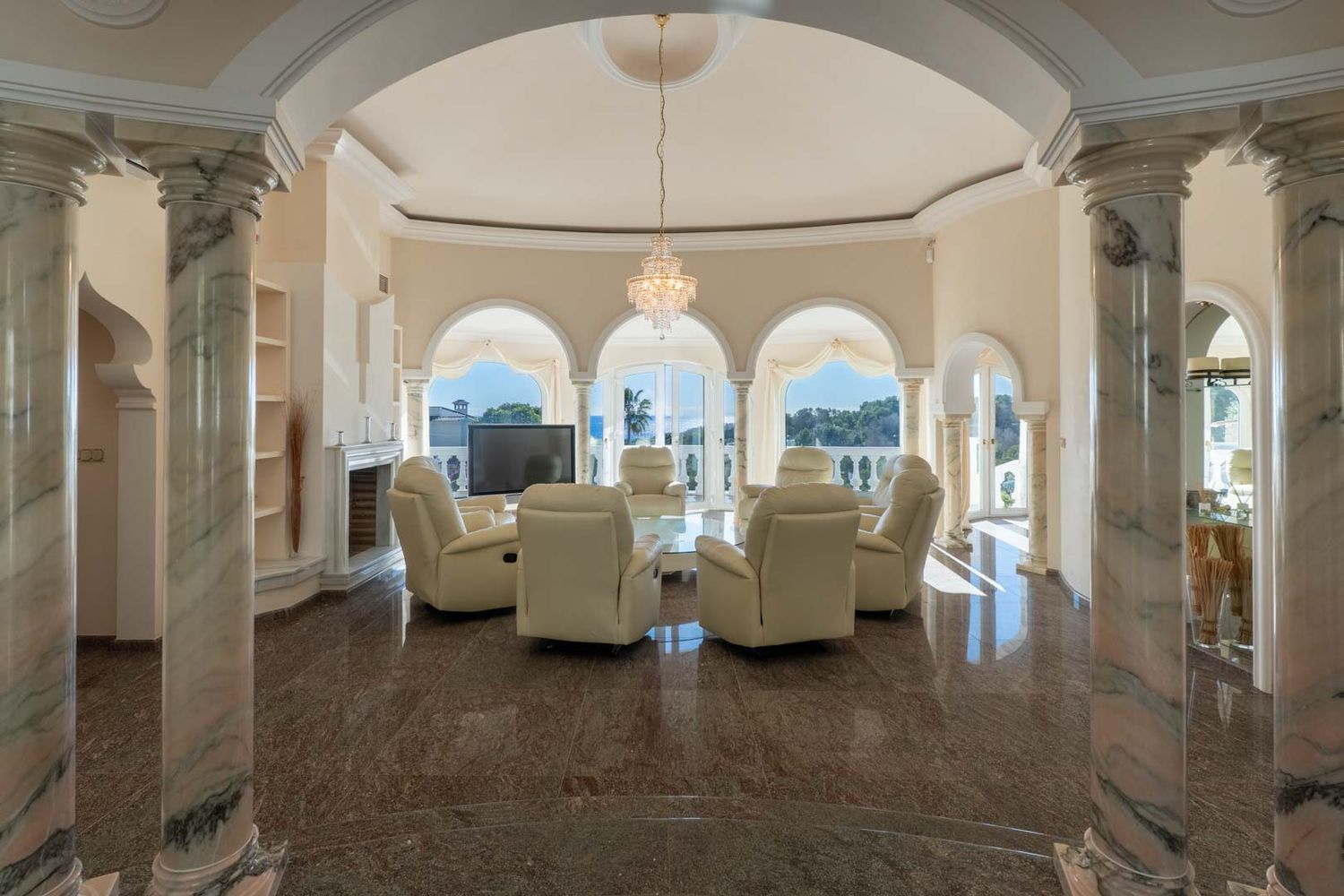 Grand living room featuring polished dark marble floors and elegant marble columns. Arched windows offer bright natural light and views, complementing the central seating area with recliners and a crystal chandelier.