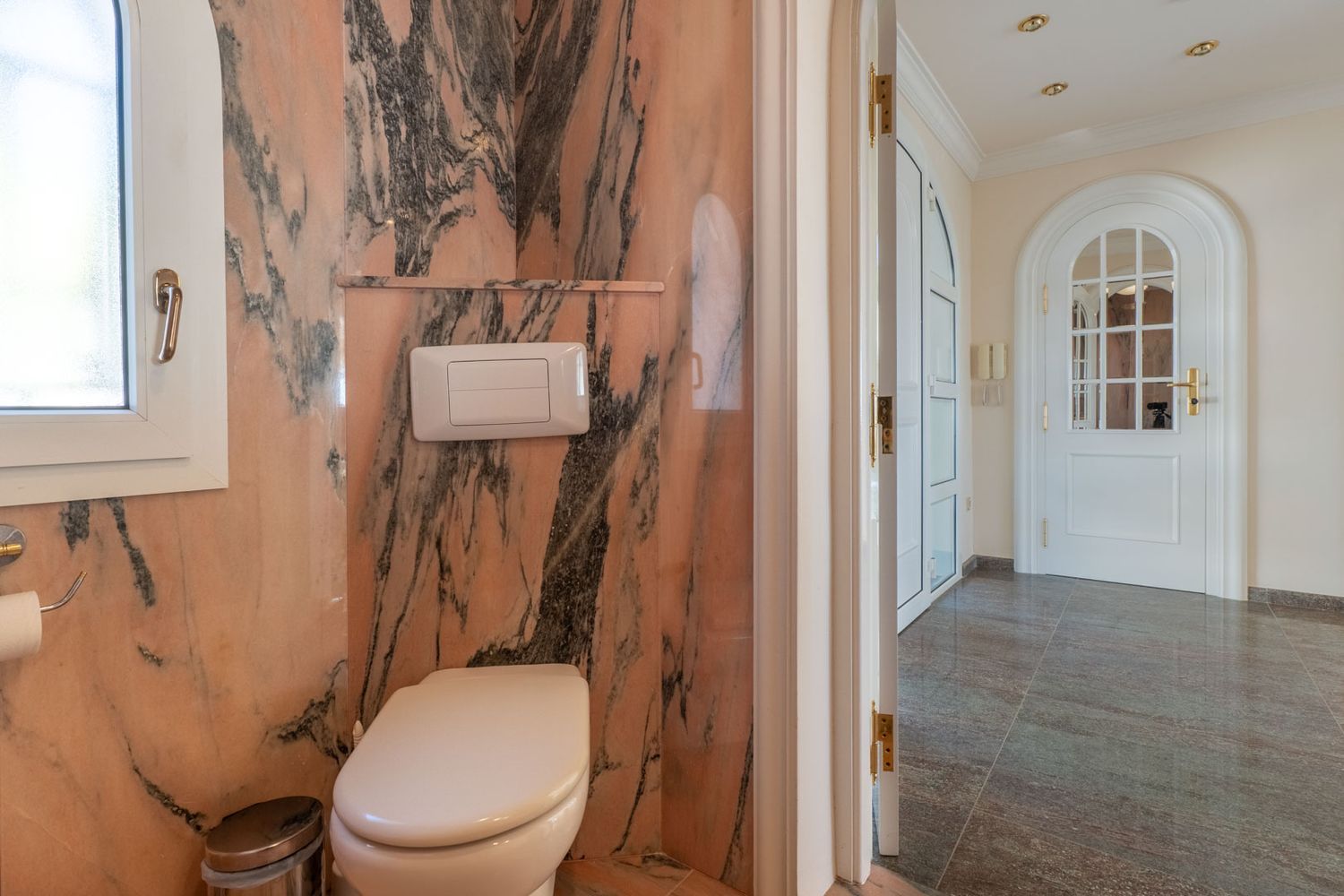 Guest toilet featuring pink veined marble walls and a window, connecting to a hallway with dark tiled flooring and an arched door.