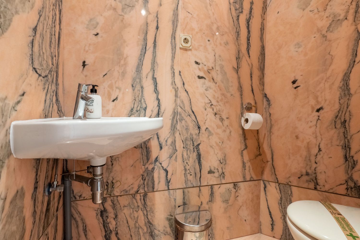 Guest toilet featuring walls and floor clad in pink veined marble. Equipped with a wall-mounted white sink, chrome faucet, and toilet.