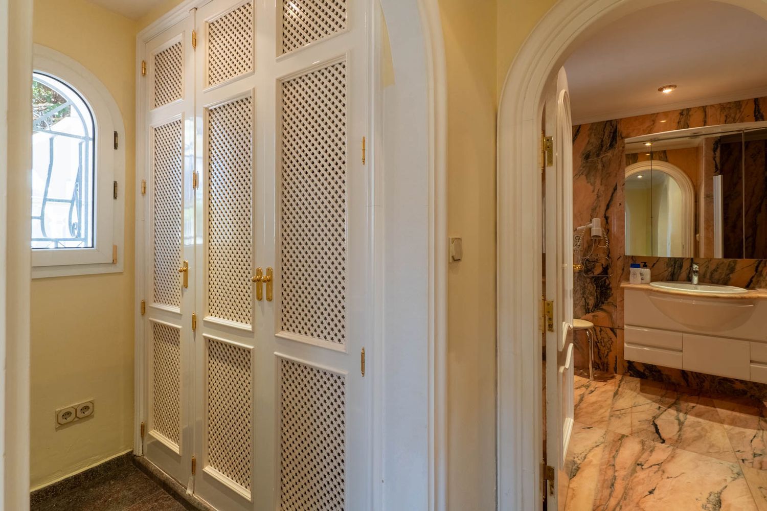 Hallway featuring white louvered built-in wardrobes with gold handles and an arched window. An arched doorway leads to a bathroom with veined marble finishes.