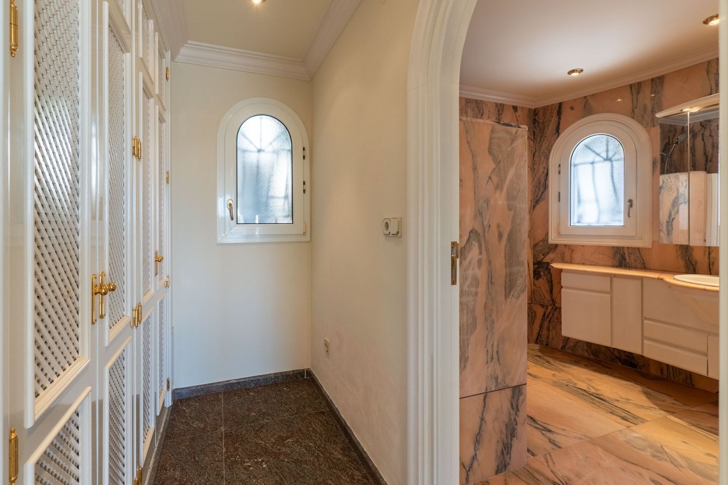 Hallway featuring white louvered wardrobes with gold handles, leading to a bathroom clad in pink marble with two arched windows.