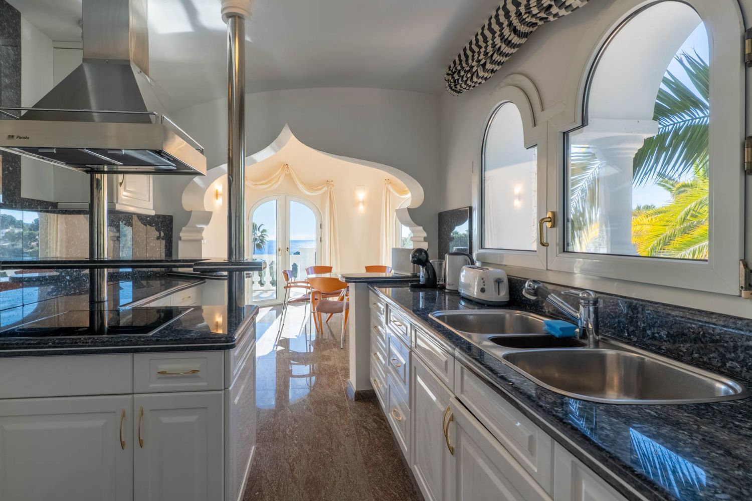 Independent kitchen featuring dark granite countertops and a double sink. Decorative arches frame the Mediterranean sea view and dining area, with polished marble flooring.