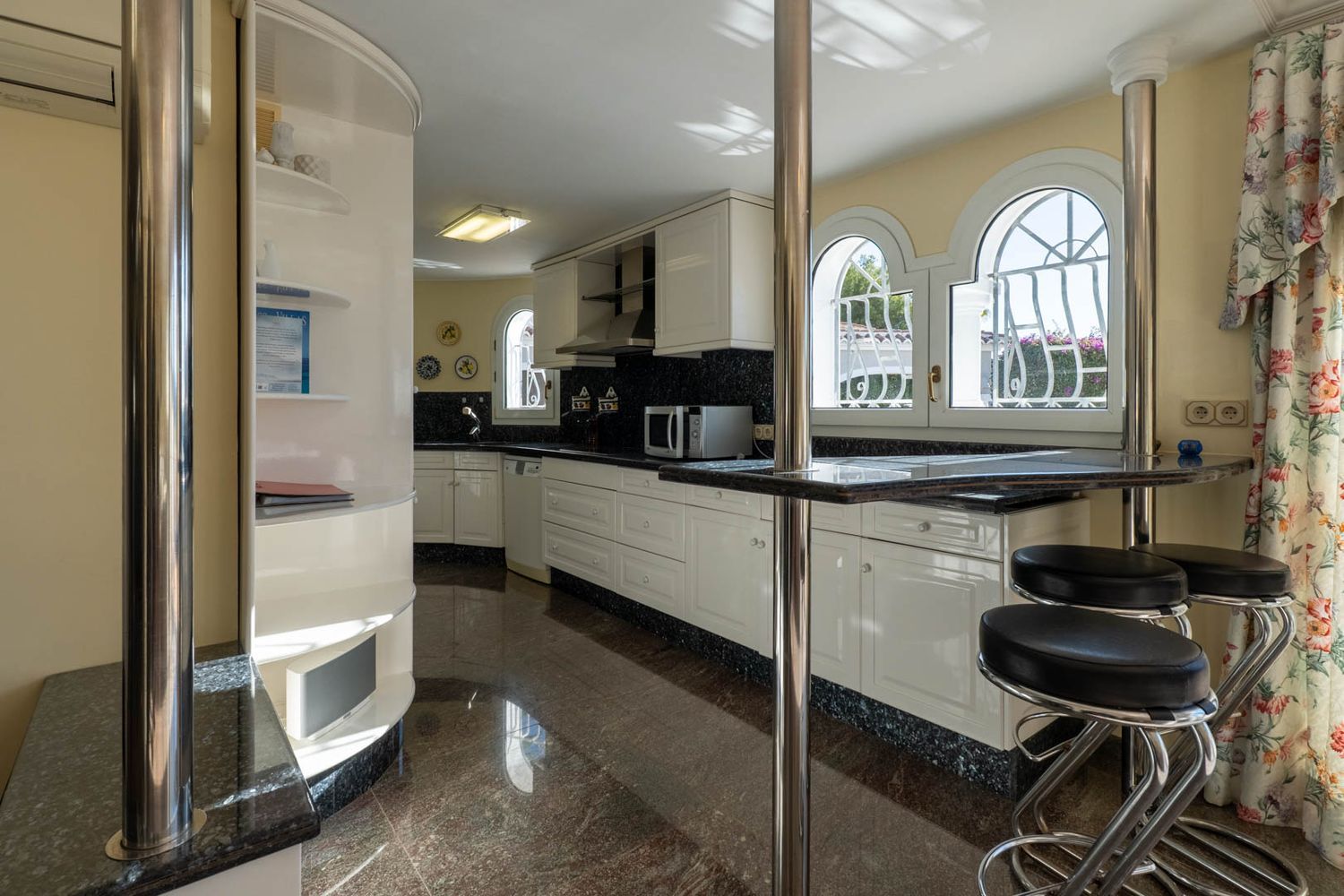 Independent kitchen featuring white cabinetry, dark granite countertops, and a breakfast bar with stools. Arched windows offer views of the exterior, complemented by a polished reddish-brown floor.